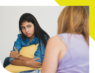 Teenager sitting with arms crossed, holding a pillow with rear view of adult looking on